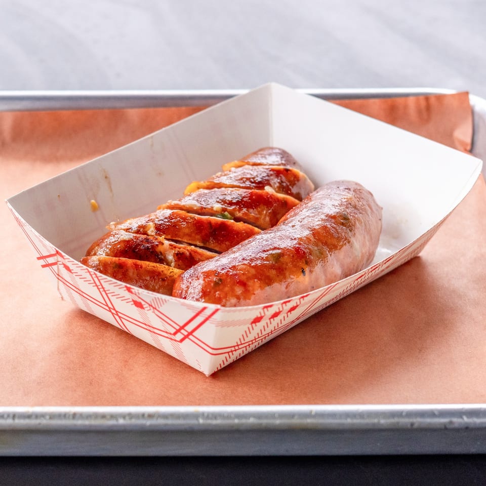 Hudson Smokehouse bx BBQ sausage, whole and sliced, in a paper tray on a metal baking sheet.