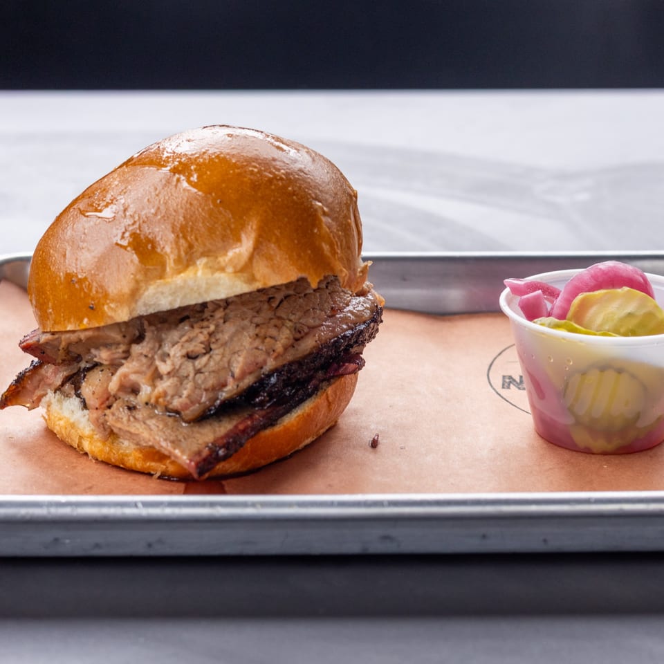 Hudson Smokehouse's brisket sandwich with a side of pickled onions and cucumbers on a metal tray.