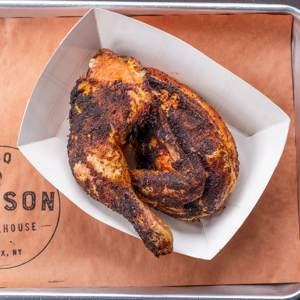 A half smoked chicken with crispy, dark skin in a white paper tray from Hudson Smokehouse.