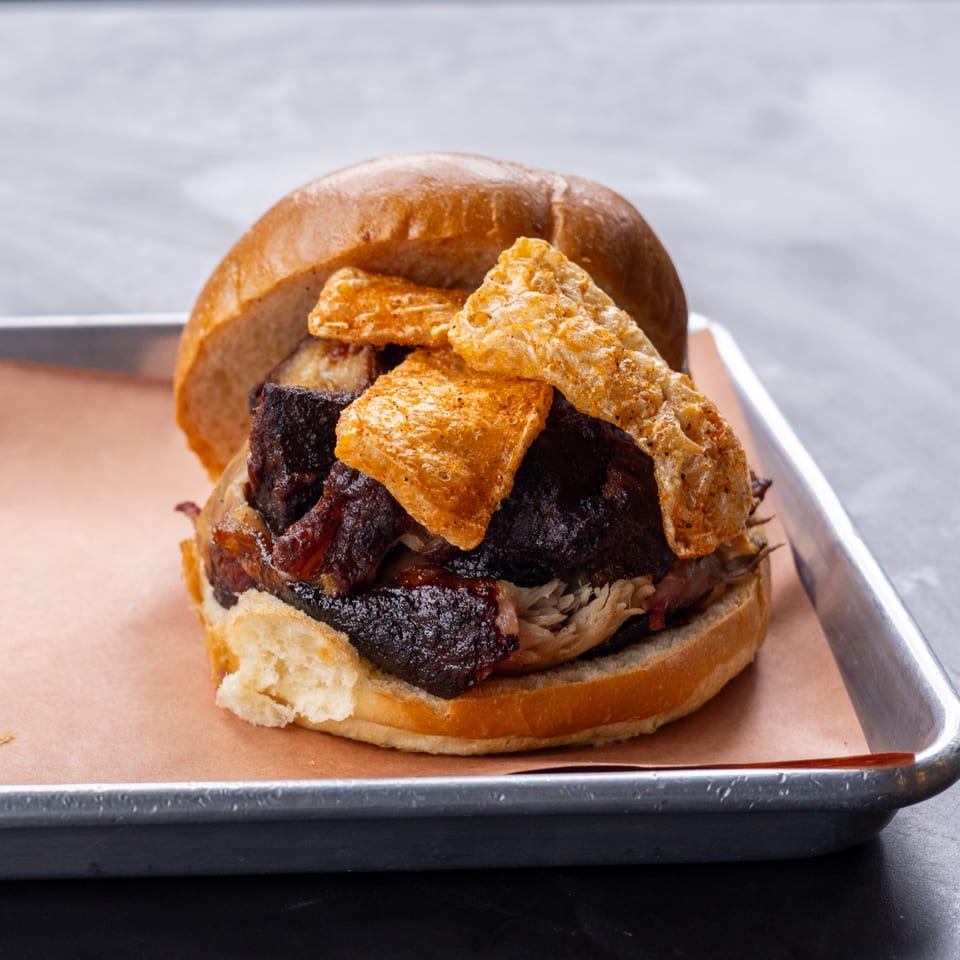Hudson Smokehouse bx BBQ sandwich with dark meat, pulled pork, and pork rinds on a bun.