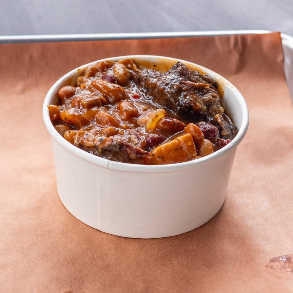 A white cup filled with Hudson Smokehouse bx baked beans and brisket on a brown paper-lined tray.