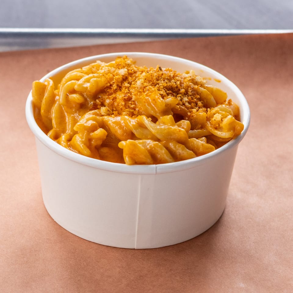 A white paper cup filled with Hudson Smokehouse bx's creamy mac and cheese, topped with breadcrumbs.