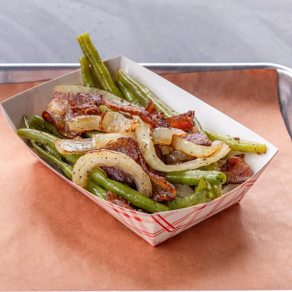 Green beans with caramelized onions and bacon bits in a paper tray, a Hudson Smokehouse bx side.